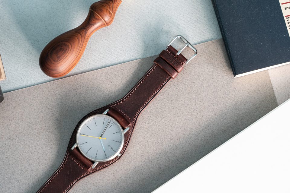Dark brown Bund strap with a pale-dial watch beside an awl and notebook.