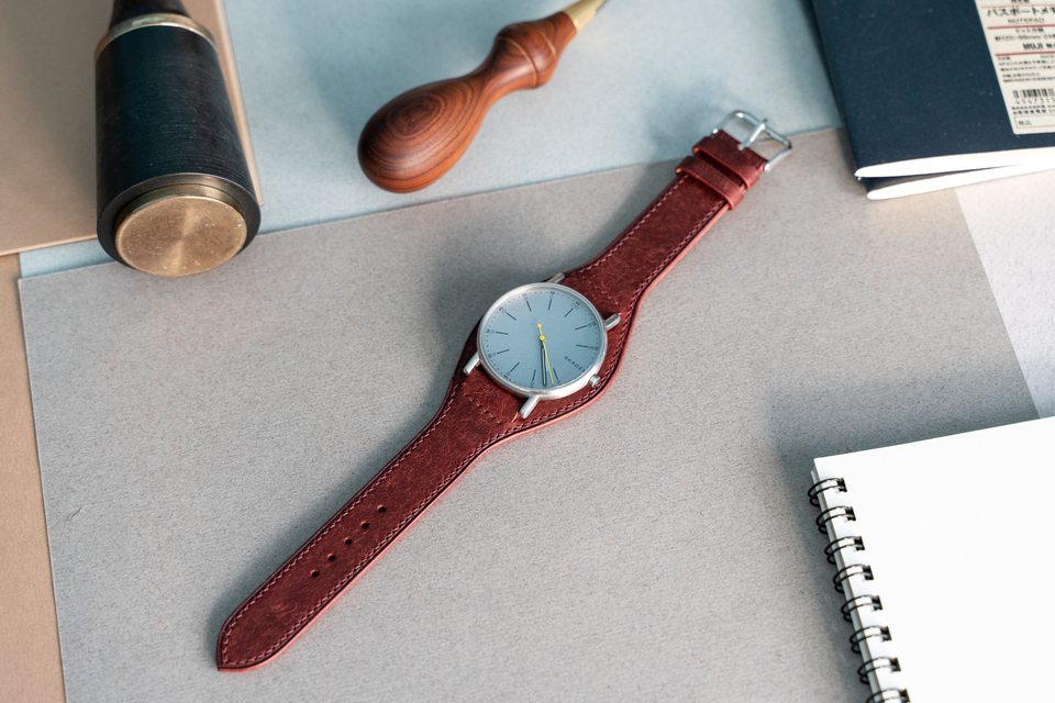 Red Bund strap with a pale-dial watch beside a mallet and awl.
