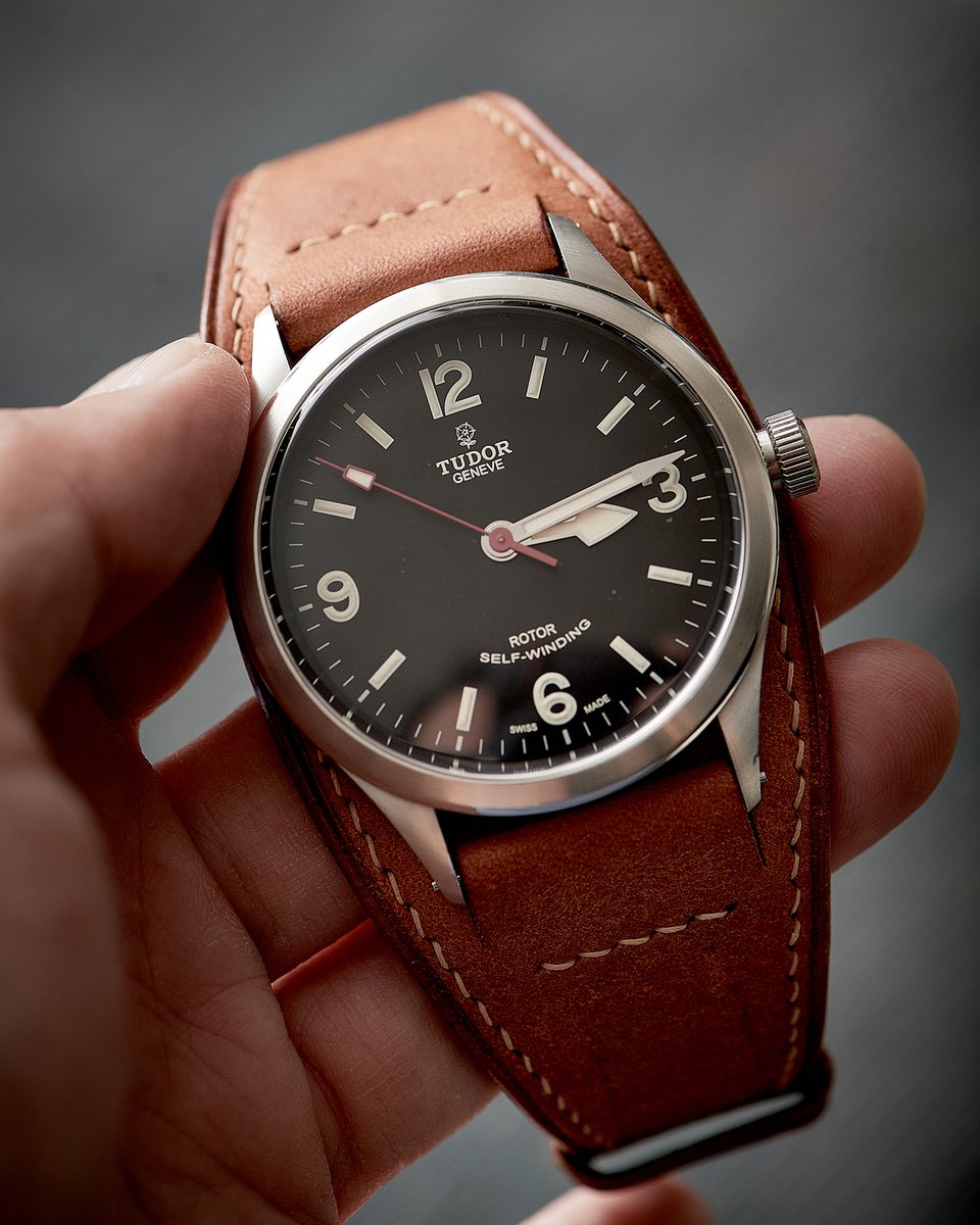 Dark-dial watch on a broad tan Bund strap, held for a close view of the leather surround.